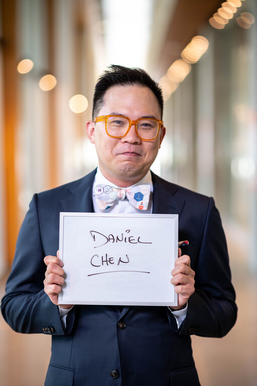 Daniel Chen in 2022, his first year at UBC and in MDS, wearing a navy suit, orange glasses, and a hex-sticker bow tie, holding a small whiteboard with his name handwritten on it, in a sunlit UBC hallway.